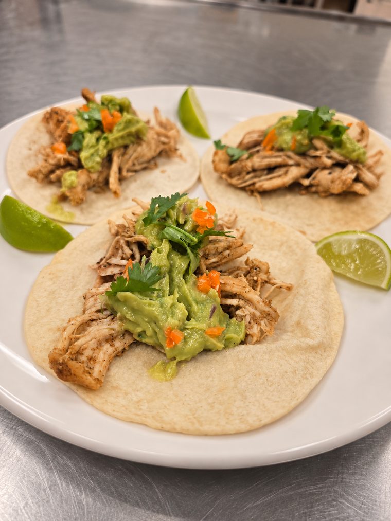 Three shredded chicken tacos topped with guacamole, minced habanero pepper and cilantro, served with lime wedges on a white plate.
