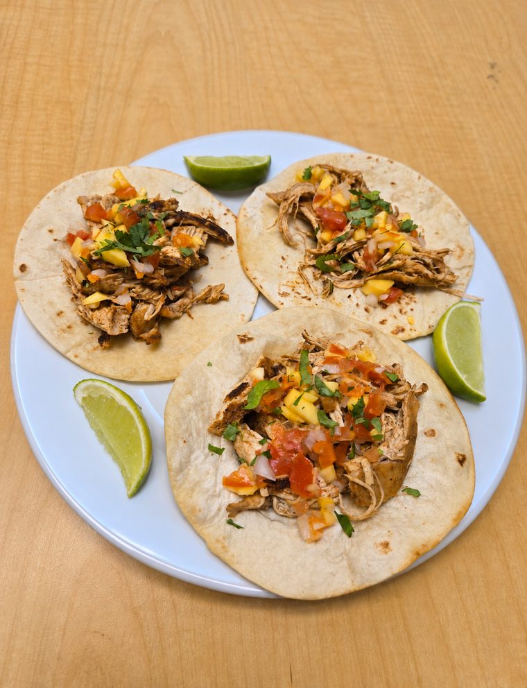 Three chicken tacos topped with mango salsa and cilantro, served with lime wedges on a white plate.