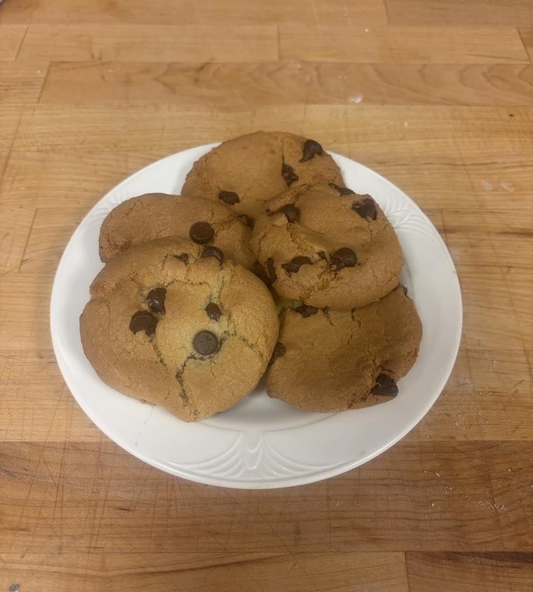 Five thick chocolate chip cookies stacked on a white plate on a wooden counter.