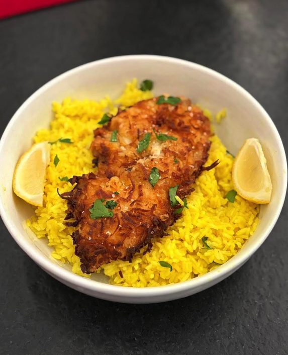 A coconut-crusted haddock fillet on a bed of yellow lemon rice in a white bowl, with chopped parsley and two lemon wedges.