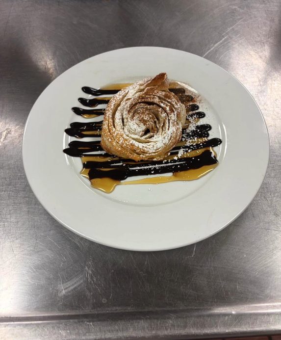 A round coiled croissant dusted with powdered sugar, plated over stripes of chocolate and caramel sauce.