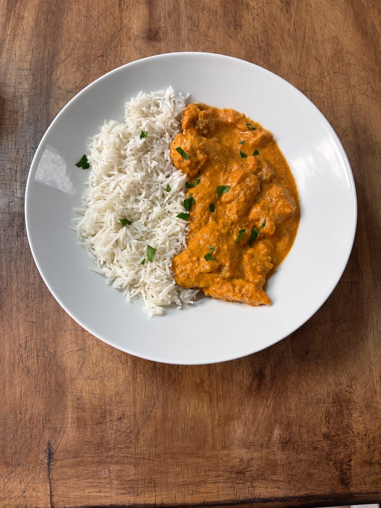 Orange curry garnished with chopped herbs beside white rice on a white plate on a wooden surface.