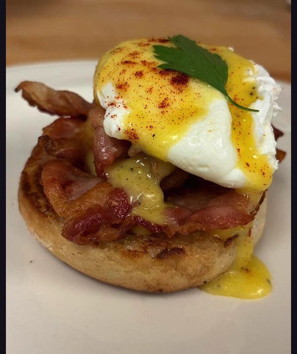 A toasted English muffin layered with bacon and a poached egg, coated in hollandaise and dusted with paprika, topped with a parsley leaf.