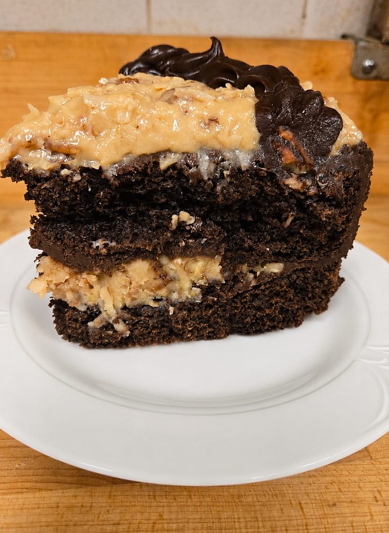 A tall slice of layered chocolate cake with coconut-pecan filling between the layers and piped chocolate frosting on top.