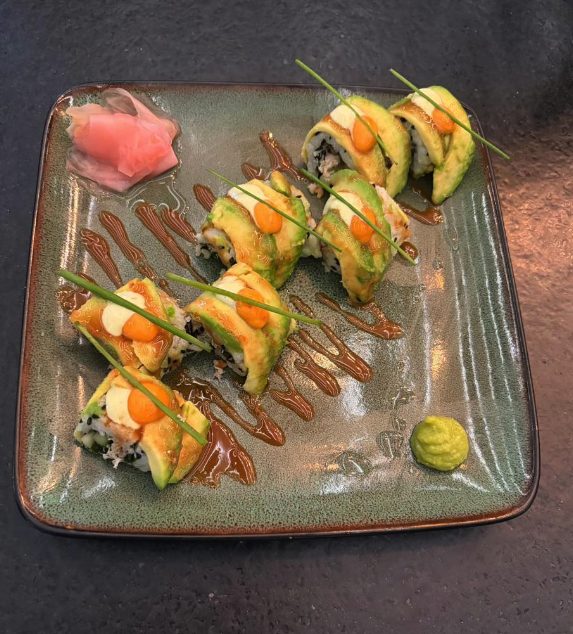 A sliced maki roll fanned across a green plate, draped in avocado with dots of orange sauce and chives, over a zigzag of dark sauce.