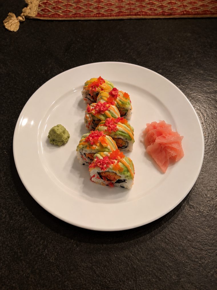 A sliced maki roll topped with avocado, orange roe, pink crunch and drizzles of orange and pale green sauce, with wasabi and pickled ginger.