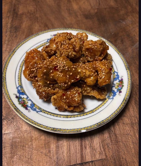 Pieces of fried chicken in a glossy orange glaze, scattered with sesame seeds and chilli flakes, on a floral-rimmed plate.