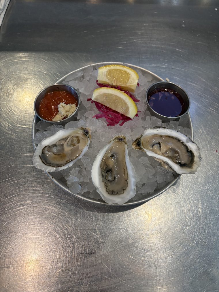 Three oysters on the half shell on crushed ice, with ramekins of cocktail sauce and mignonette, lemon wedges and pickled red cabbage.