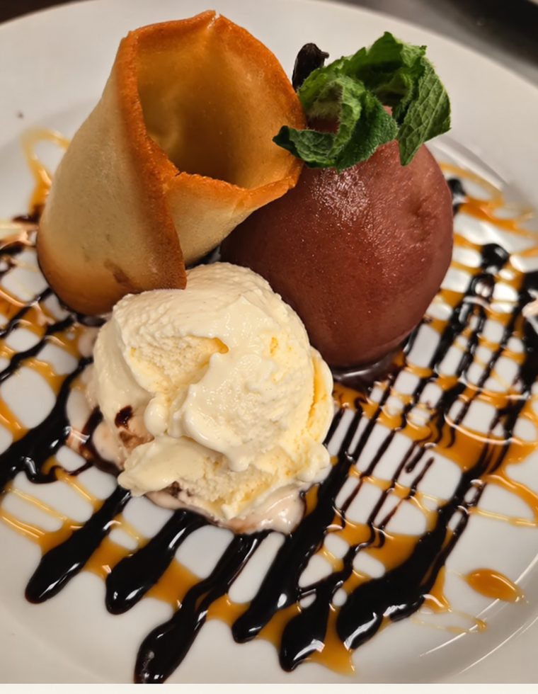 A poached pear with a mint sprig beside a scoop of vanilla ice cream and a curled tuile, over chocolate and caramel sauce.