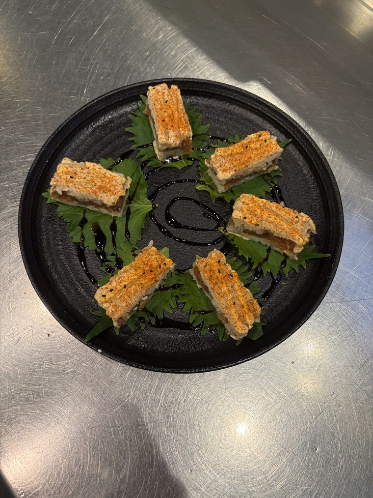Six rectangles of pressed sushi on shiso leaves arranged in a circle on a black plate, each dusted with chilli and sesame, over a swirl of dark glaze.