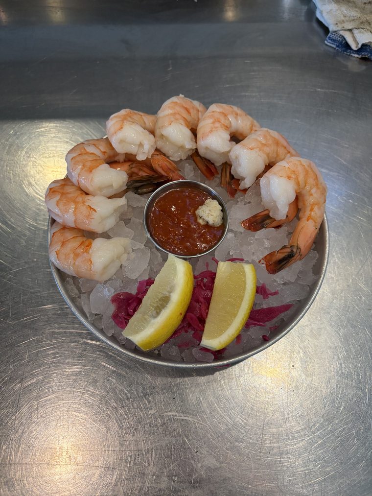 Eight poached shrimp arranged in a ring on crushed ice around a ramekin of cocktail sauce topped with horseradish, with two lemon wedges.