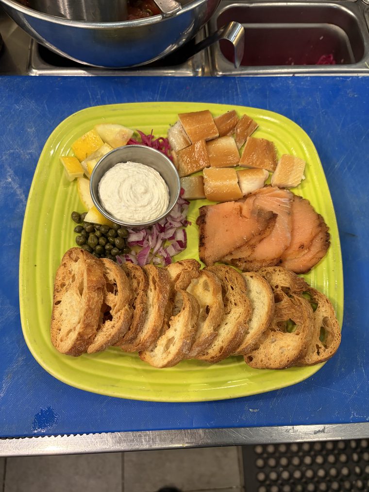 A platter of smoked salmon slices and cubes of smoked white fish with a cup of whipped spread, capers, diced red onion, lemon and a fan of grilled crostini.