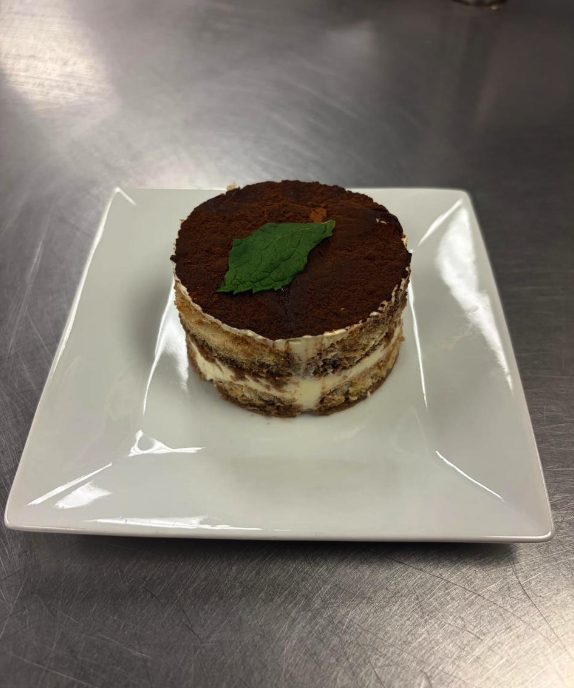 A round slice of tiramisu dusted with cocoa and topped with a mint leaf on a square white plate.