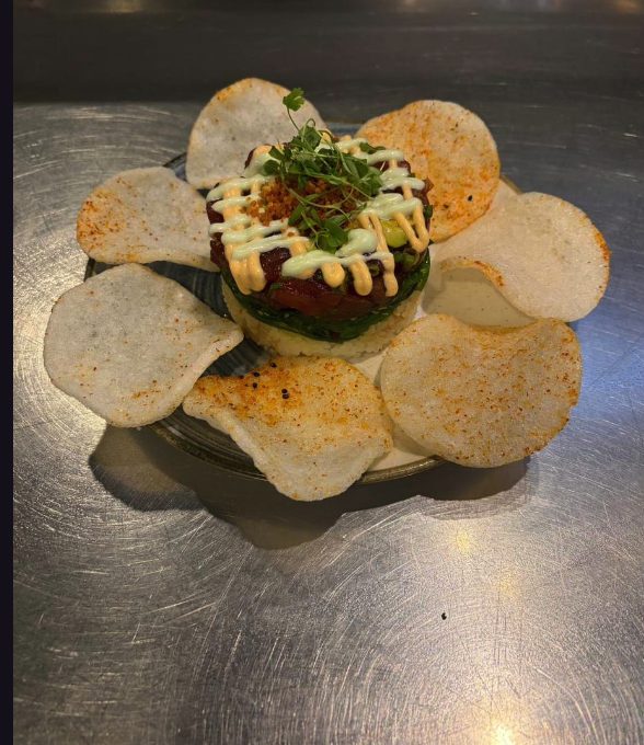 A stacked round of rice, avocado and diced tuna topped with microgreens and drizzled aioli, ringed by pale chips dusted with chilli.